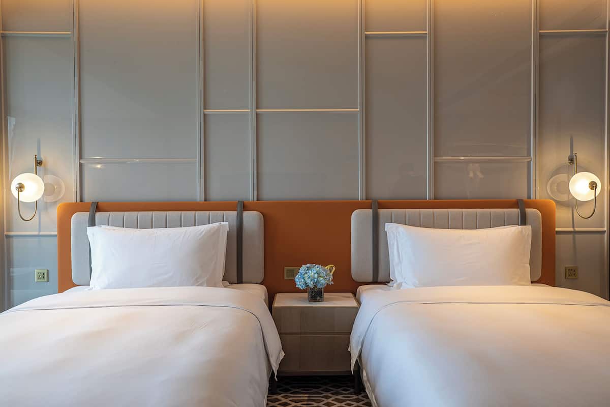A hotel room with two beds, a nightstand with a vase of flowers, and wall-mounted lamps.
