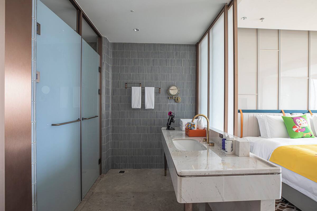 A hotel room with a bathroom, a bed with a yellow bedspread, and a nightstand with toiletries.