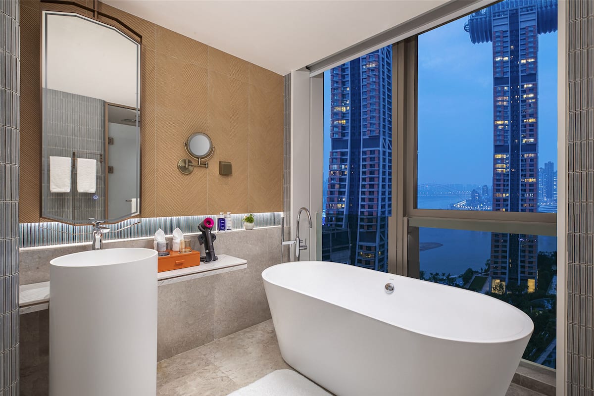 A modern bathroom with two sinks, a large bathtub, and a view of tall buildings.