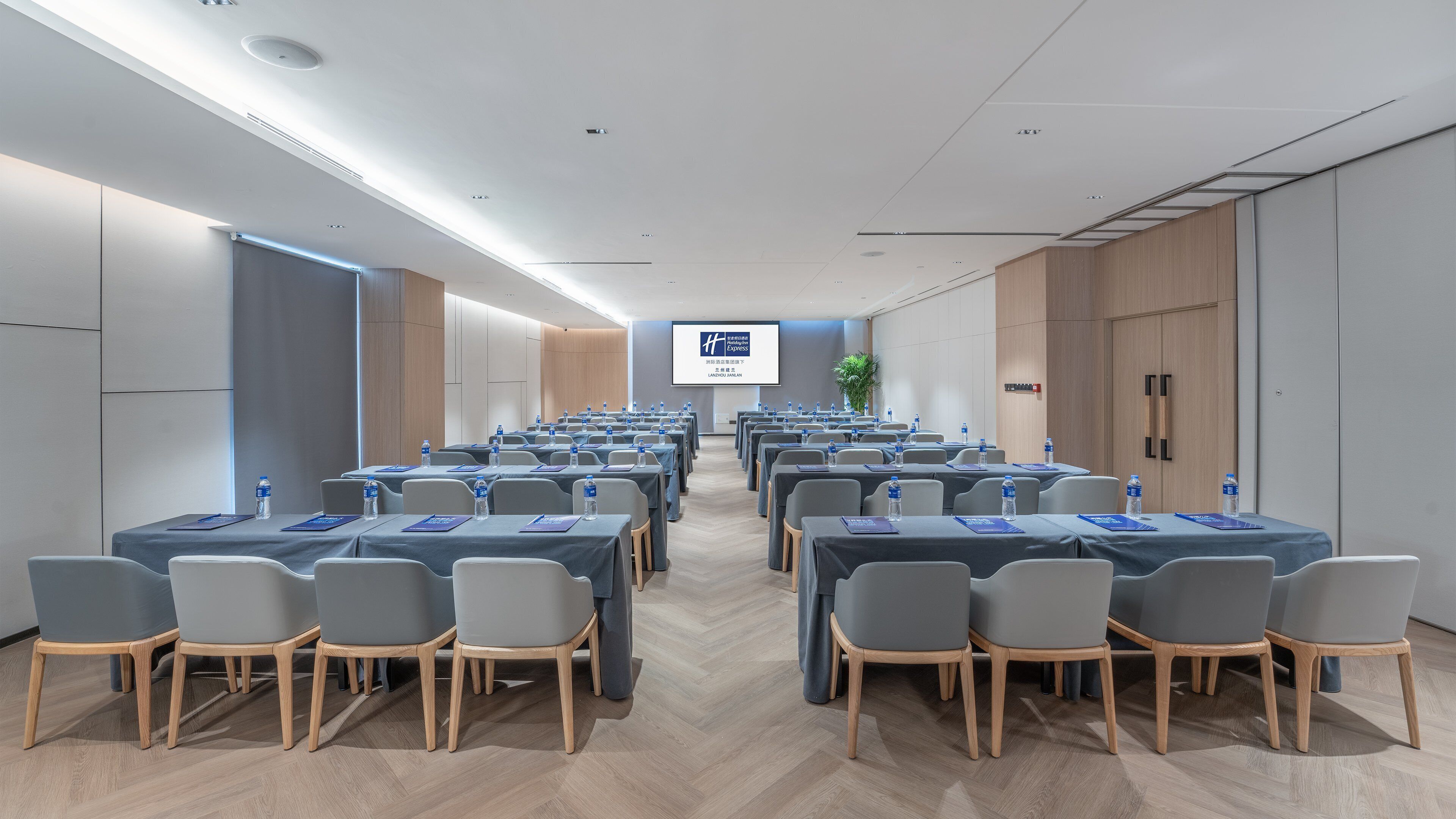 A conference room with rows of tables and chairs, a large screen at the front, and a wooden floor.