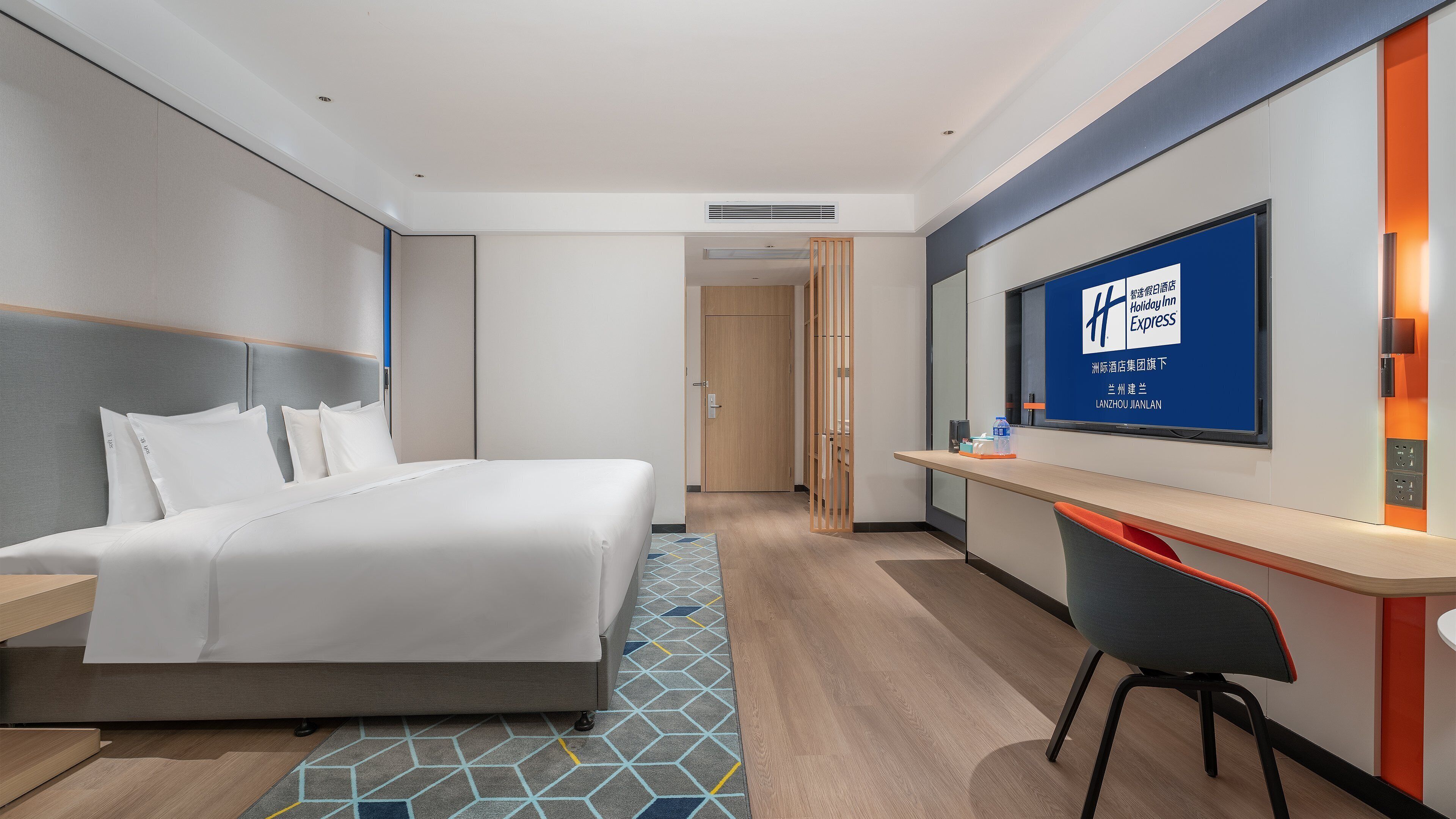 A hotel room with a bed, a desk, a chair, a TV, and a door.