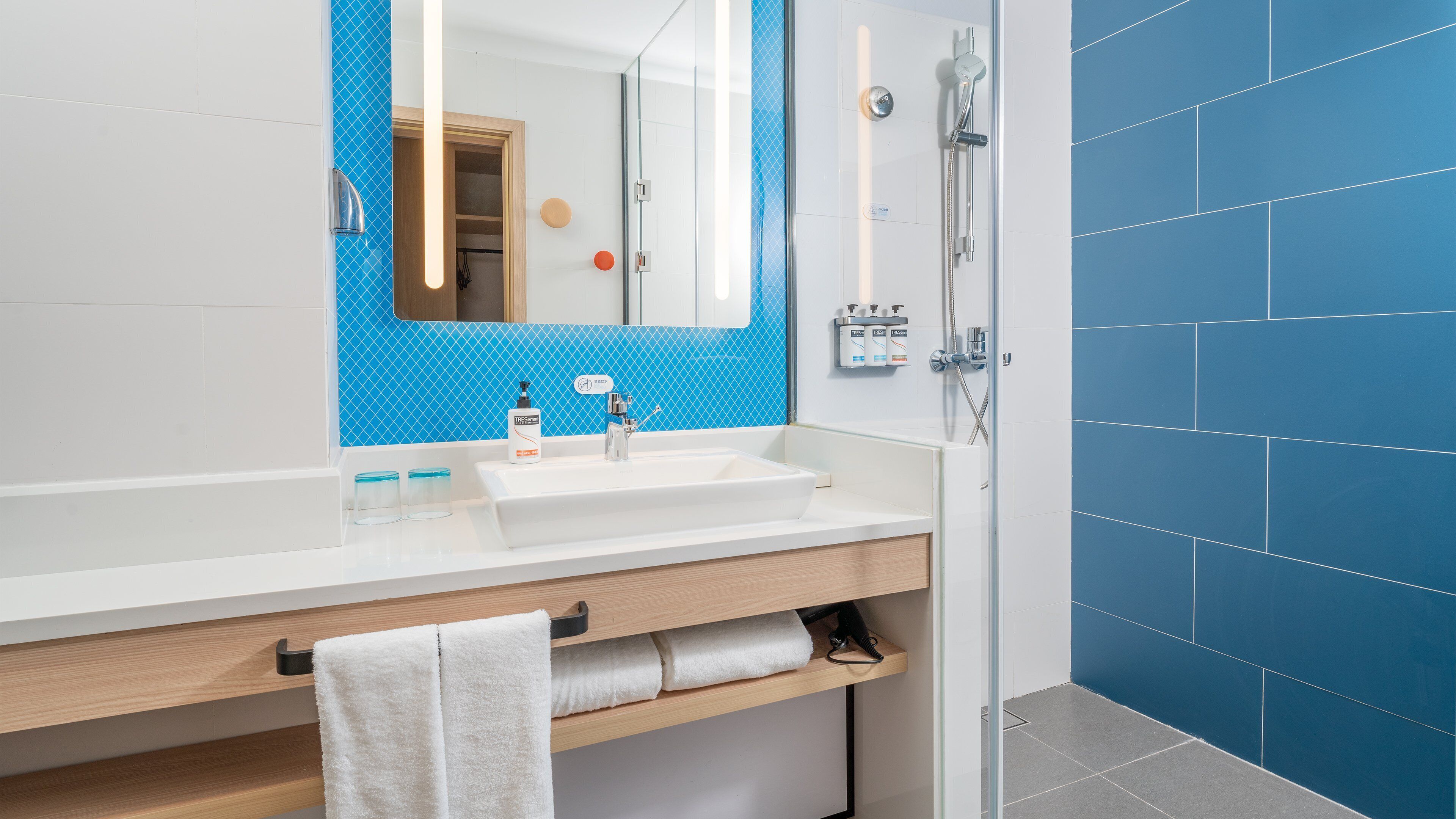 Een moderne badkamer met een wit wastafelmeubel, een douche met blauwe tegels en een houten opbergkast.