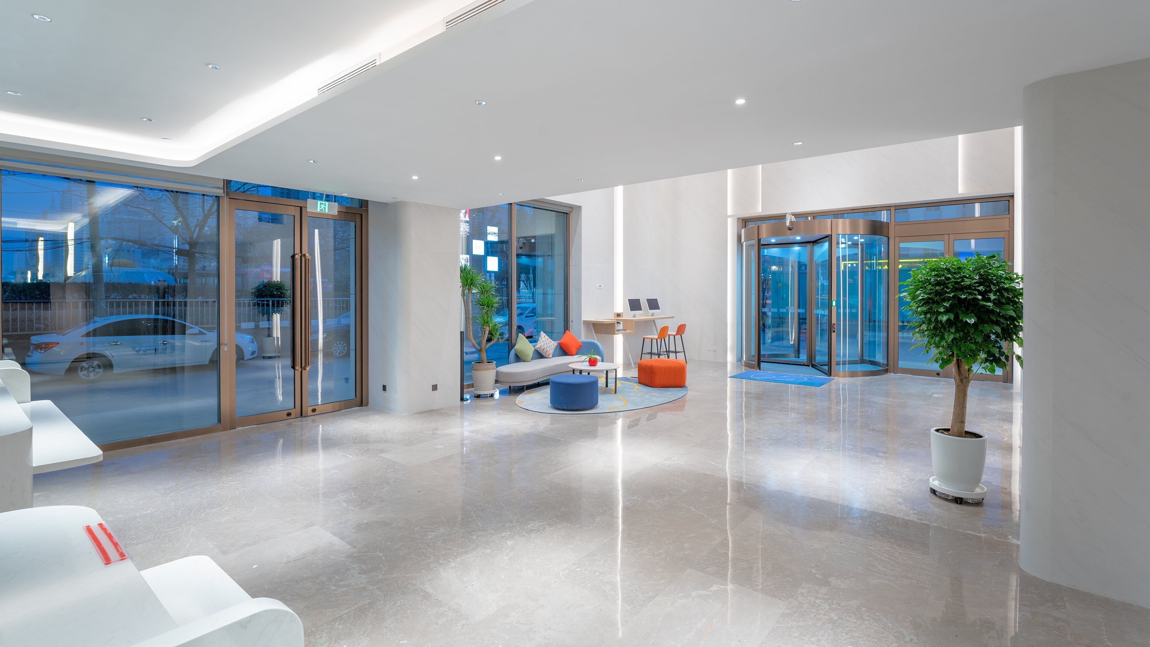 A modern lobby with glass doors, a seating area with a sofa and ottoman, a small table, and a potted plant.