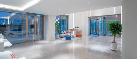A modern lobby with glass doors, a seating area with a sofa and ottoman, a small table, and a potted plant.