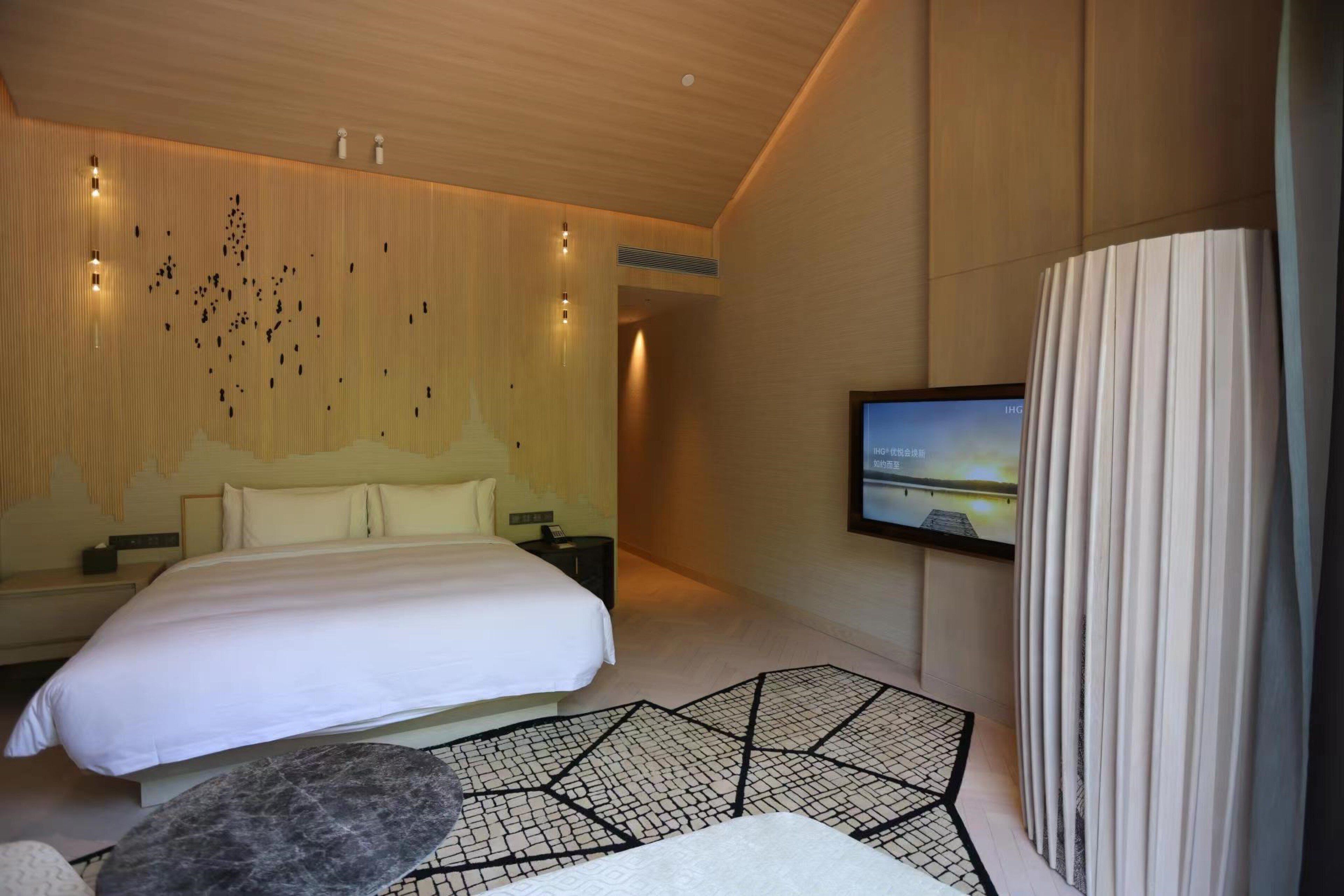 A modern hotel room with a large bed, a flat-screen TV mounted on the wall, and a unique geometric-patterned rug.