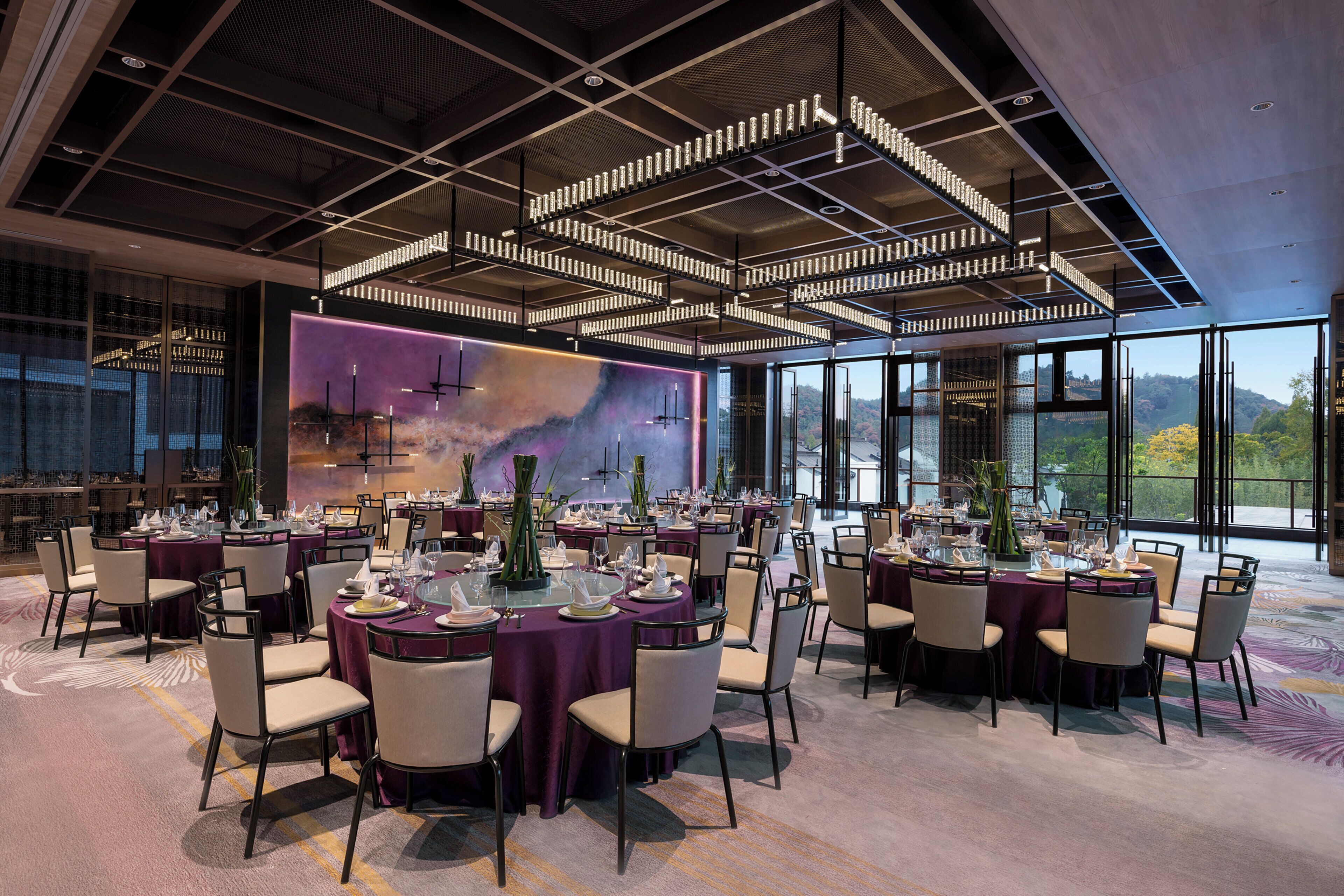 A spacious dining area with round tables set for a meal, large windows offering a view of the outdoors, and a modern ceiling design with exposed beams and lighting fixtures.