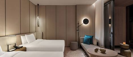 A modern hotel room with two beds, a bedside table with a lamp, a small round table with a kettle, and a large window with curtains.