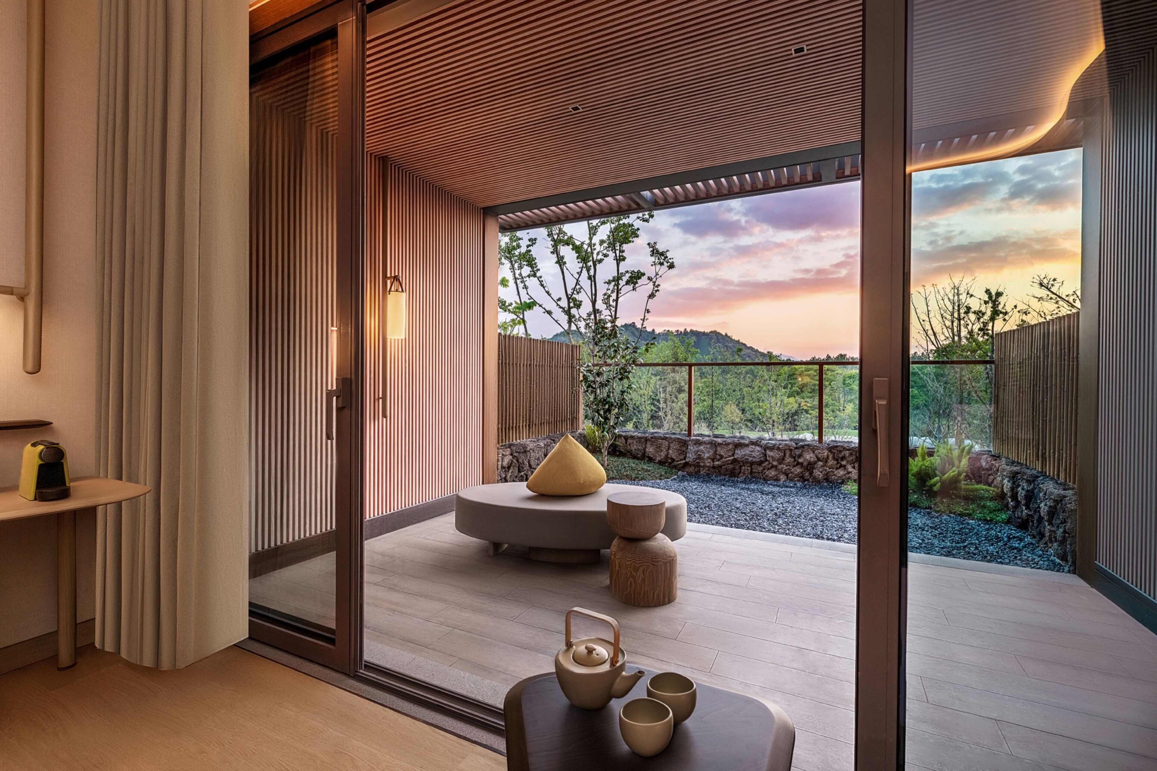 A modern interior with a sliding glass door leading to a balcony with a scenic view of hills and a sunset.