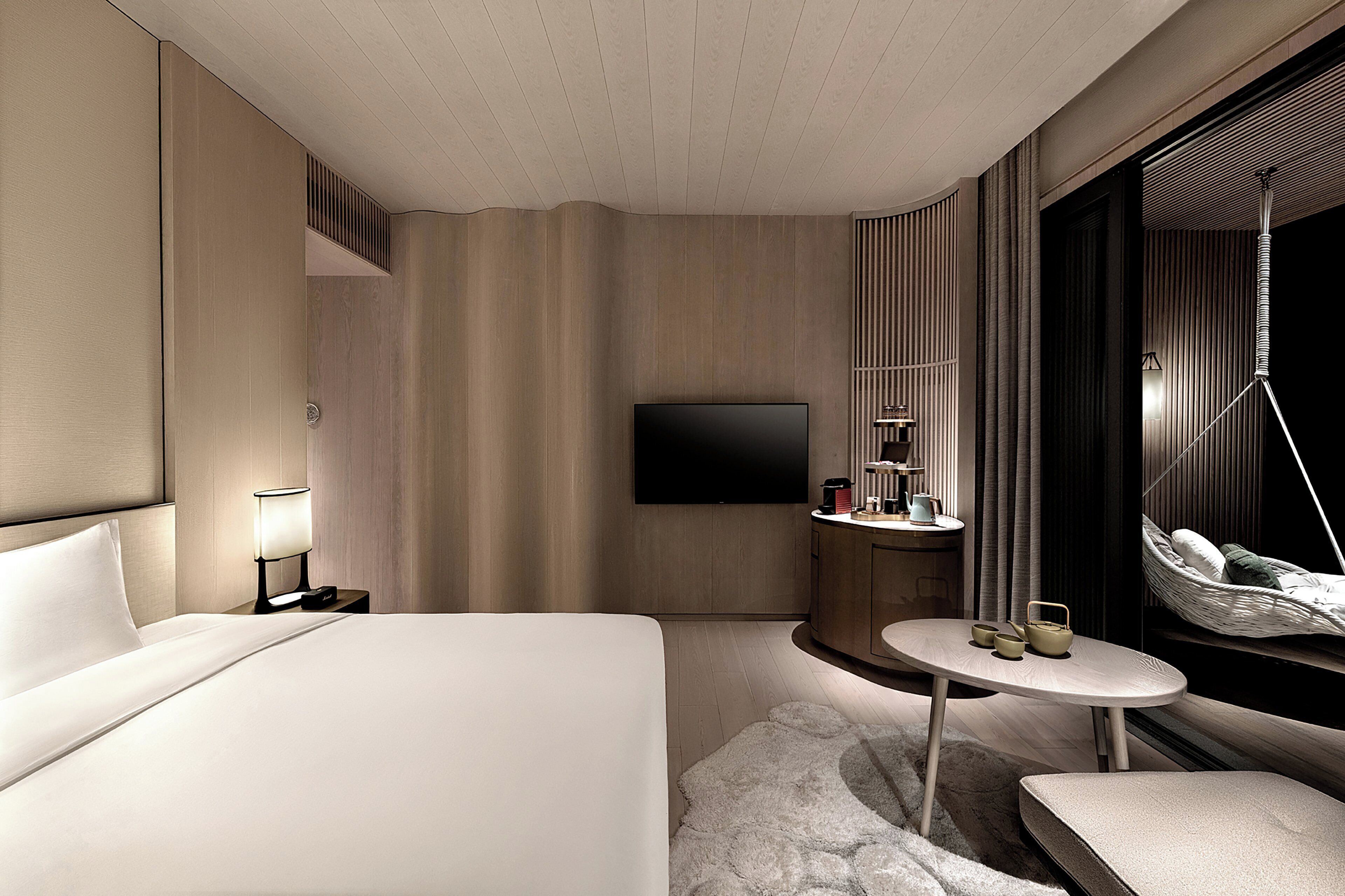 A modern hotel room with a large bed, a flat-screen TV, a small round table with a lamp, and a view of the city through a large window.