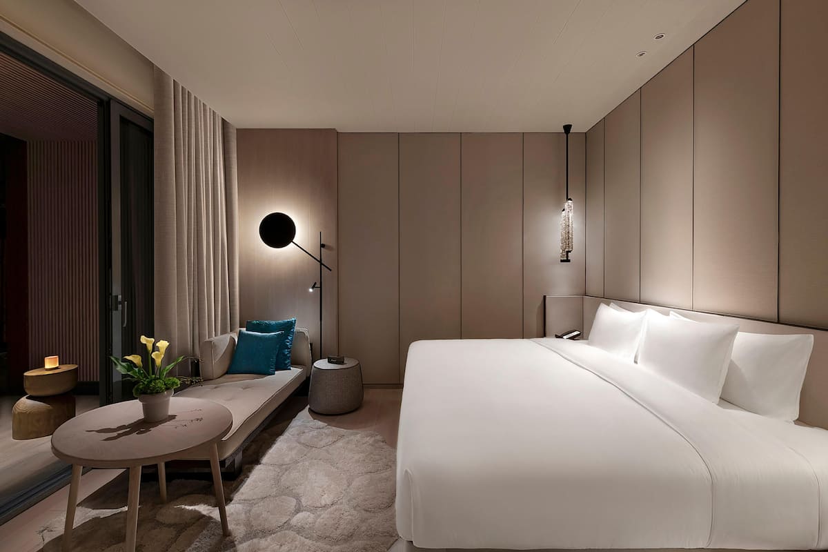 Une chambre d’hôtel moderne avec un grand lit, une table ronde agrémentée d’une plante, un banc et une petite table d’appoint avec une bougie.