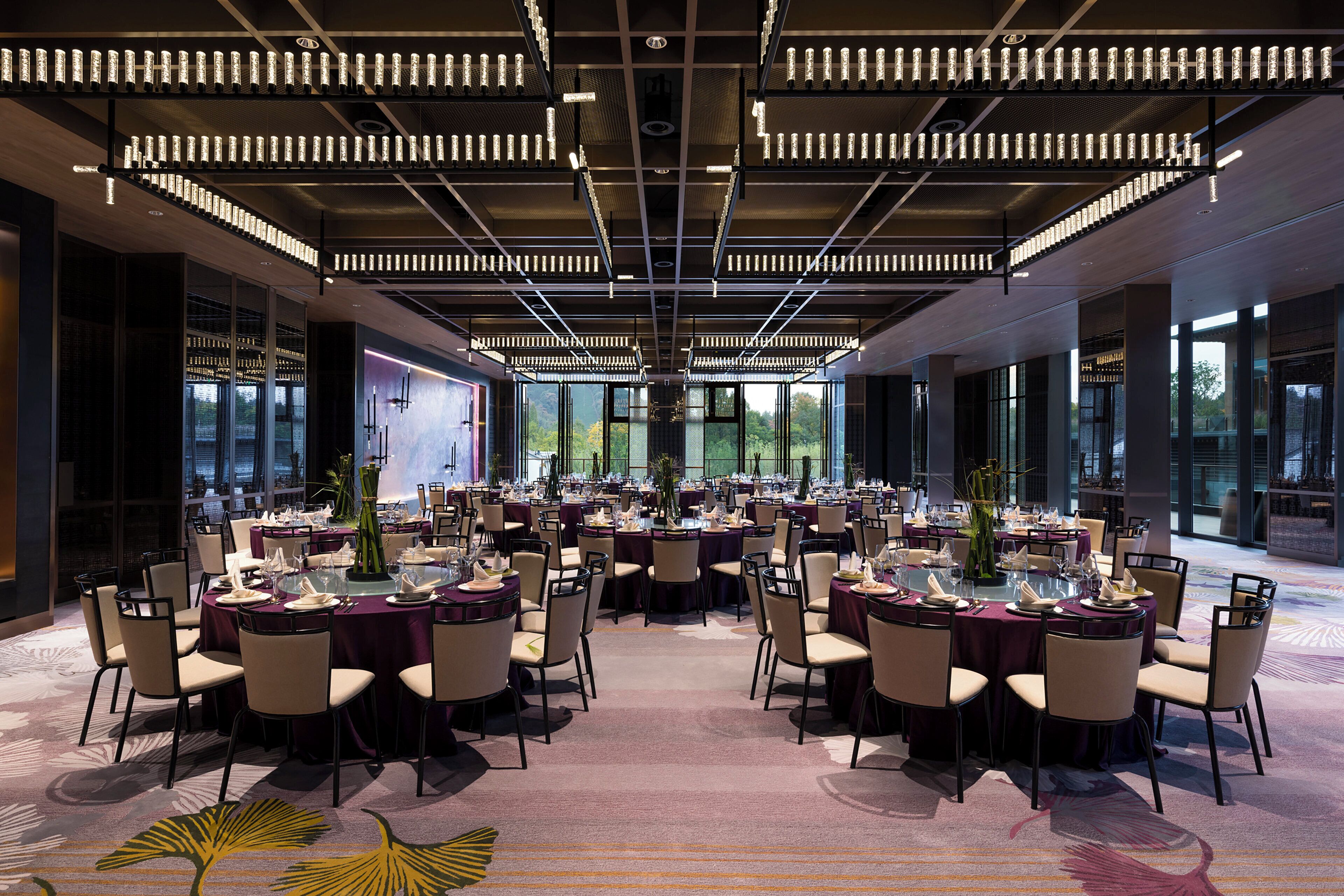 Une salle de banquet spacieuse, avec des tables rondes disposées pour le repas, de grandes fenêtres offrant une vue panoramique et un design de plafond moderne.
