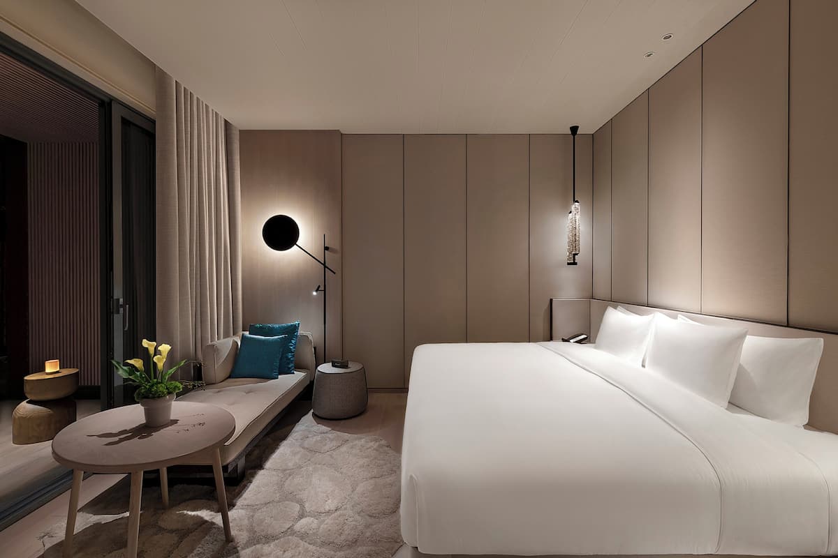 Une chambre d’hôtel moderne avec un grand lit, une table ronde agrémentée d’une plante, un banc et une table de chevet avec une lampe.