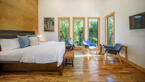 Un dormitorio con una cama de madera, un banco y una silla. Hay ventanas amplias con vista a árboles.