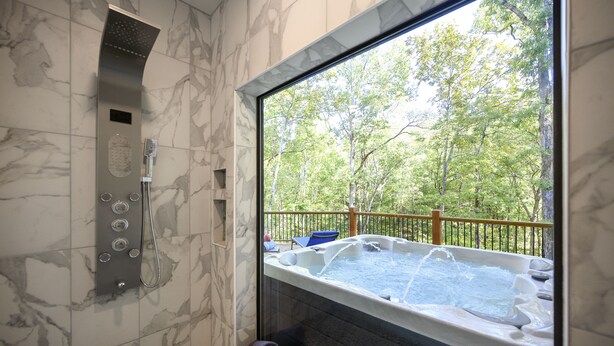 Un baño con bañera de hidromasaje, ducha y vista a un bosque a través de un ventanal grande.