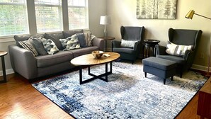 A living room with a grey sofa, two armchairs, a coffee table, and a rug. There are large windows with blinds, a floor lamp, and a painting on the wall. - Spectacular 3 BDR Townhouse Near Downtown ATL, GSU (Atlanta)