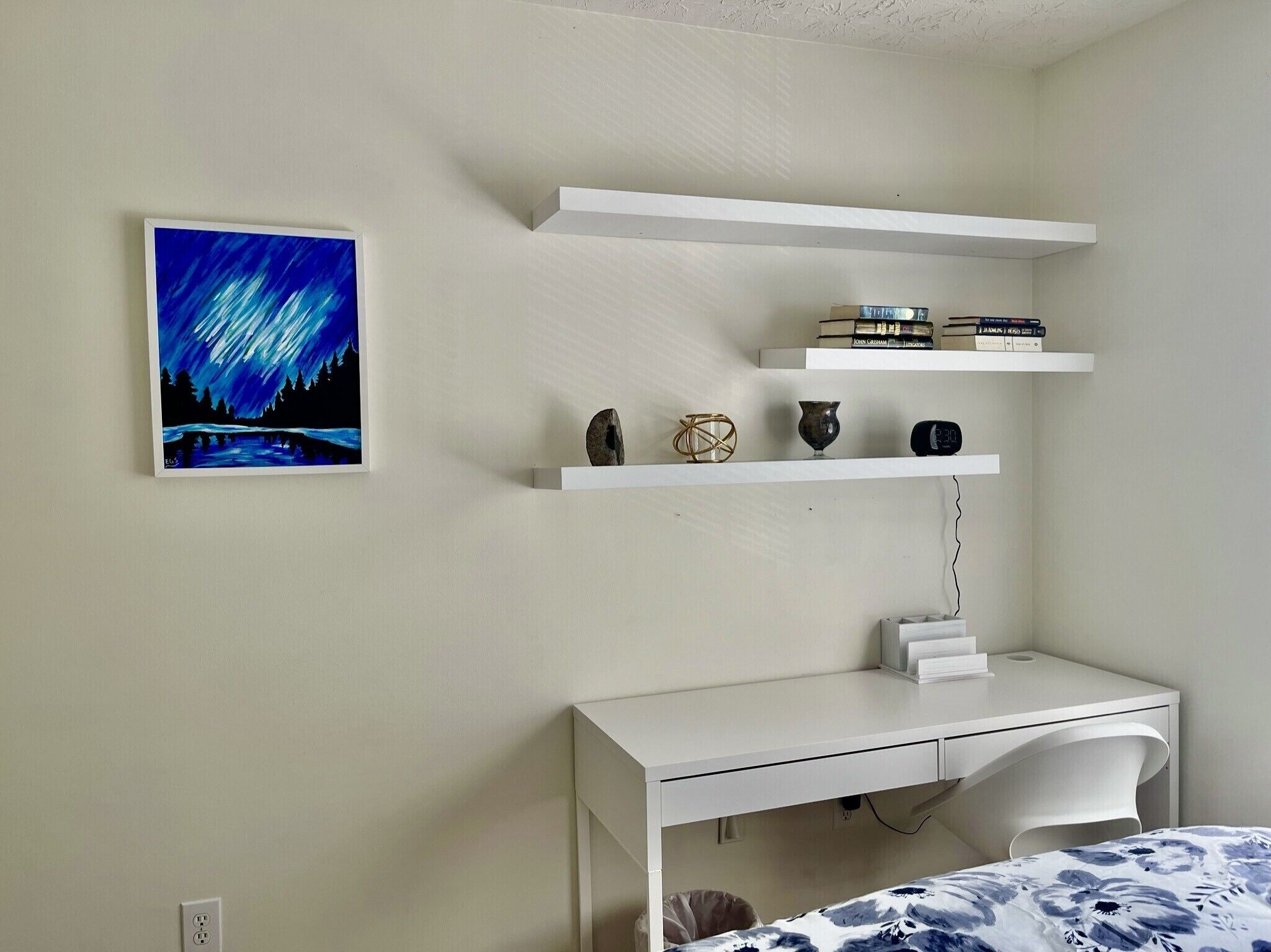 A bedroom with a white desk, a bed with a blue and white floral bedspread, and two white shelves with books and decorative items.