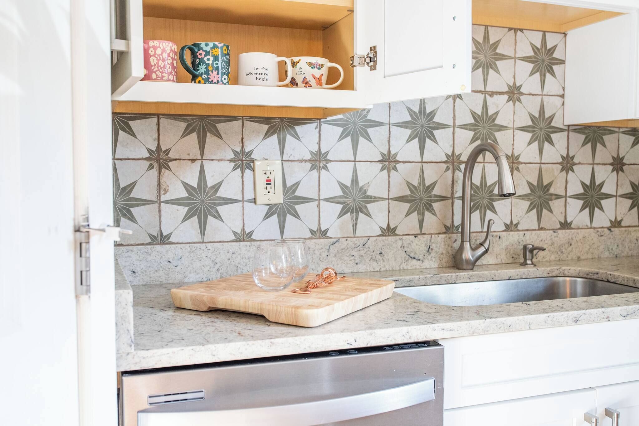 A kitchen with a granite countertop, a wooden cutting board, a stainless steel sink, and a backsplash with a geometric tile pattern.