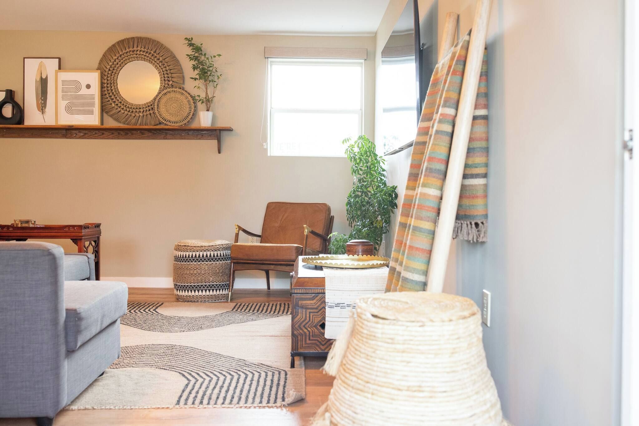 Una sala de estar con sofá, sillón y una mesa de centro. Hay objetos decorativos en una estantería, un espejo grande y una ventana con persianas.