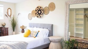 Een slaapkamer met een grijs gestoffeerd bed, witte beddengoed en decoratieve wandkunst van geweven materiaal.