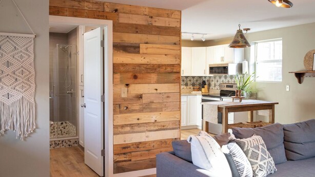 Een moderne keuken met houten details, een eethoek met een tafel en stoelen, en een grijze bank met gekleurde kussens.