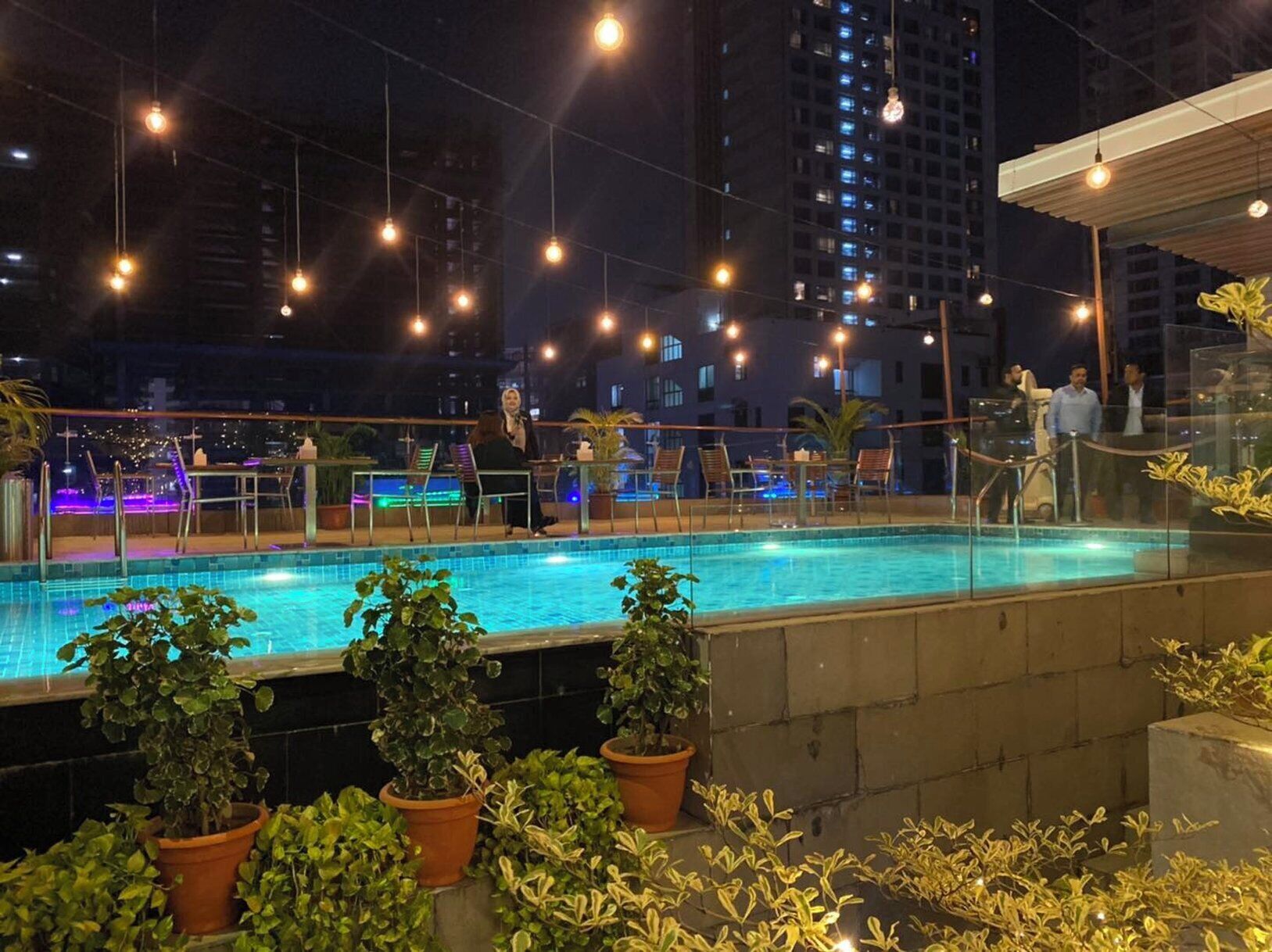 A rooftop pool area with a view of the city at night, surrounded by potted plants and outdoor seating.