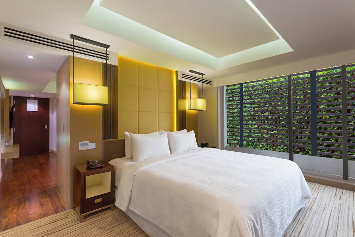 Hotelkamer met een groot bed, nachtlampjes, een nachtkastje, een wandairconditioner en een raam met uitzicht op groen.