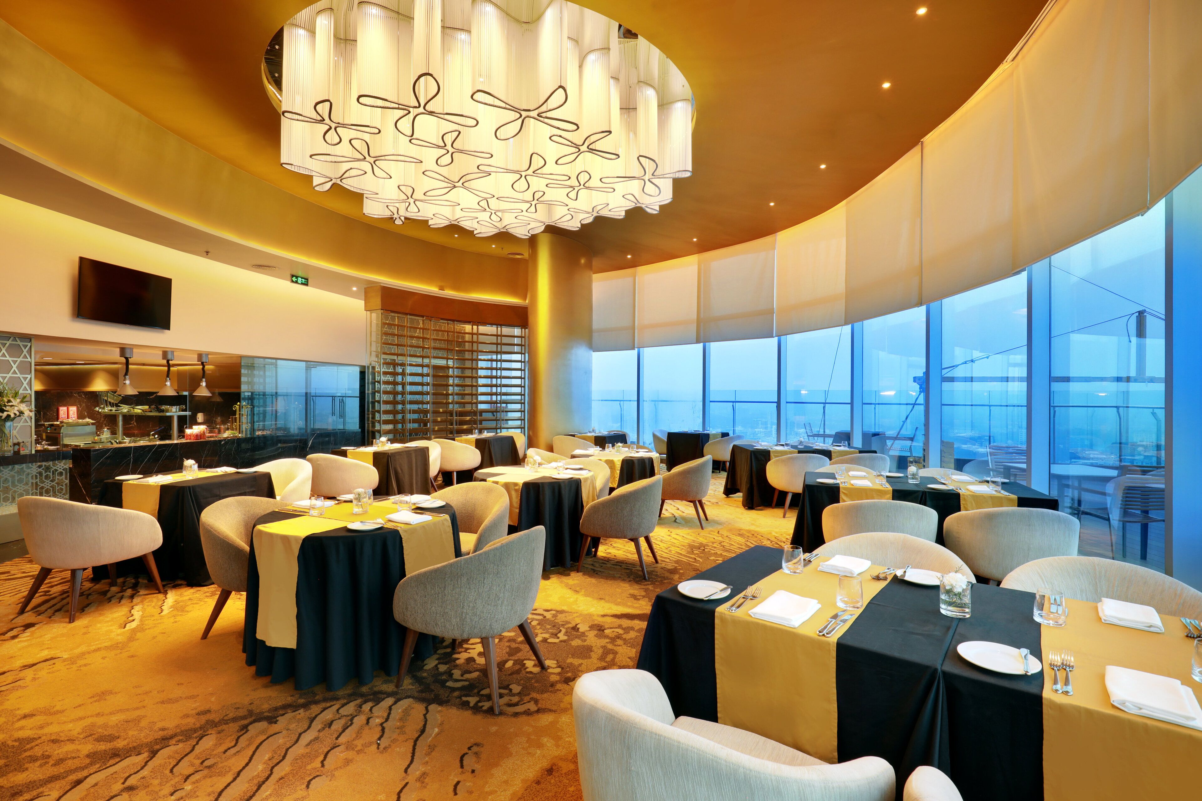 A spacious dining area with multiple tables and chairs, a modern chandelier, and large windows offering a view.