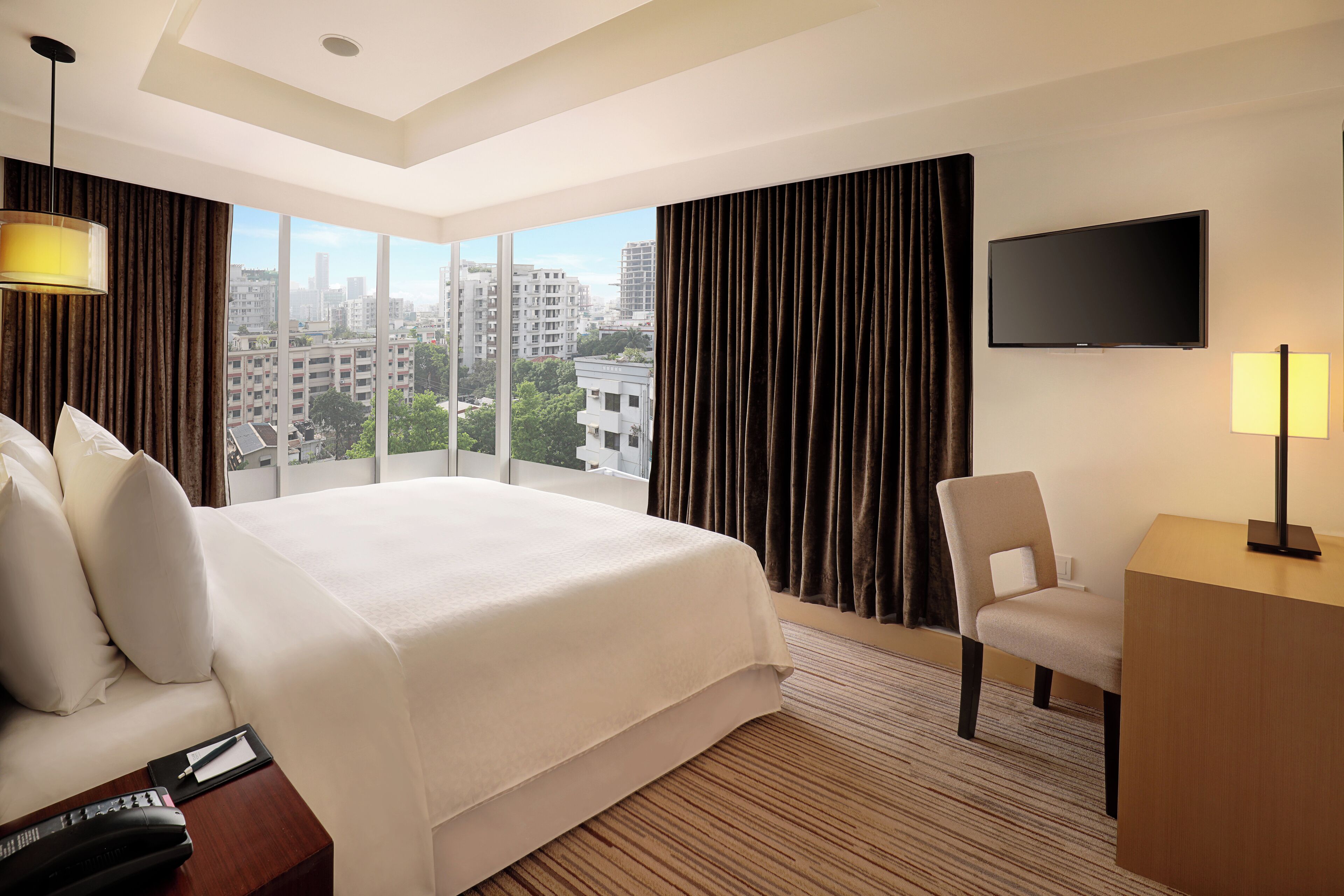 A hotel room with a large bed, a desk, a chair, a TV, and a city view through large windows.