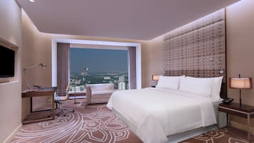 A hotel room with a large bed, a desk with a chair, a TV, and a view of the cityscape.