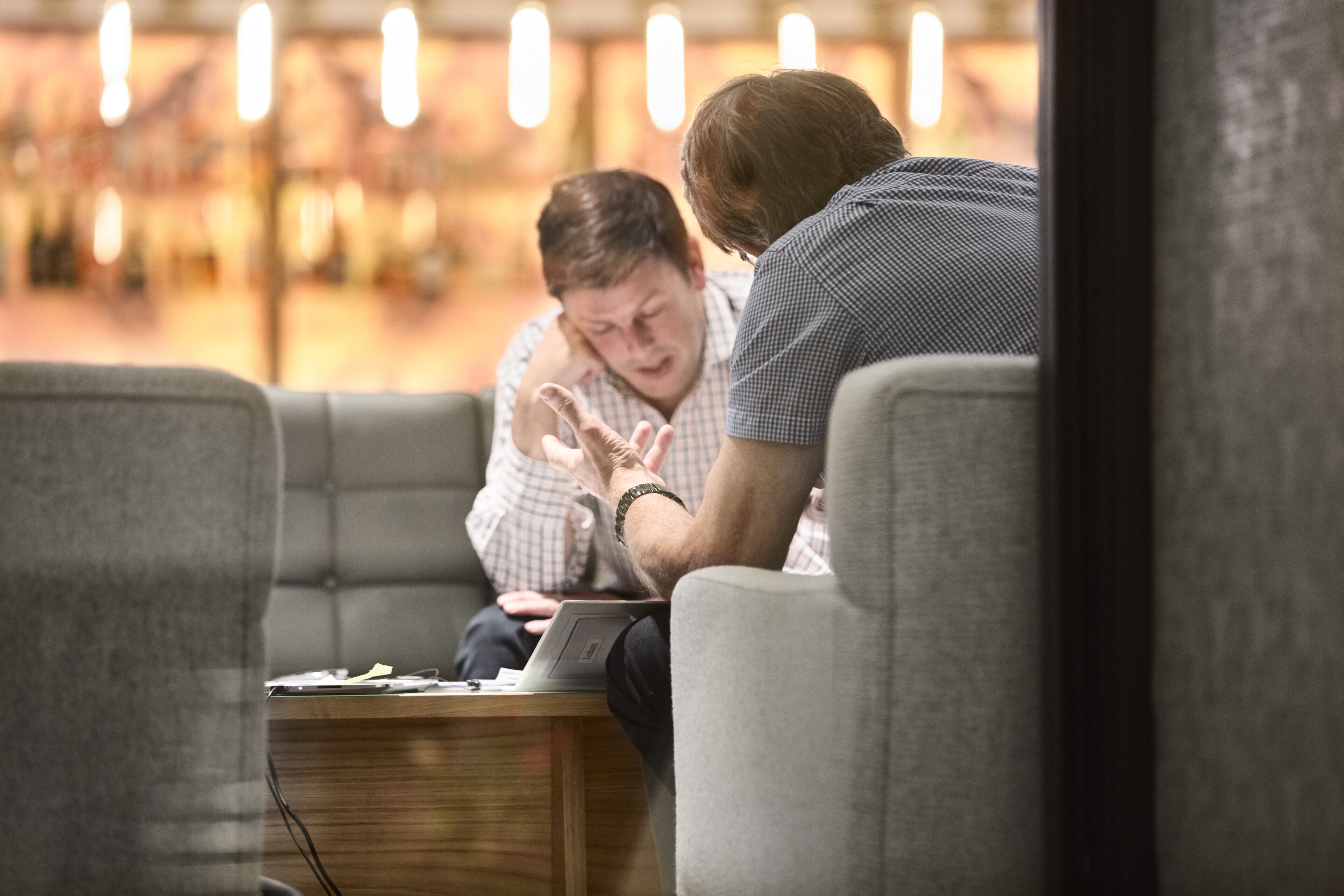 A person sitting on a couch with a laptop, another person leaning over, and a table with papers.
