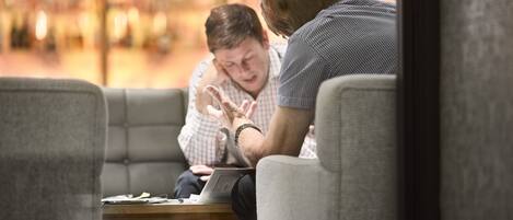 A person sitting on a couch with a laptop, another person leaning over, and a table with papers.
