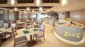 A modern restaurant with multiple dining tables and chairs, a central buffet area with various food items, and a kitchen in the background.