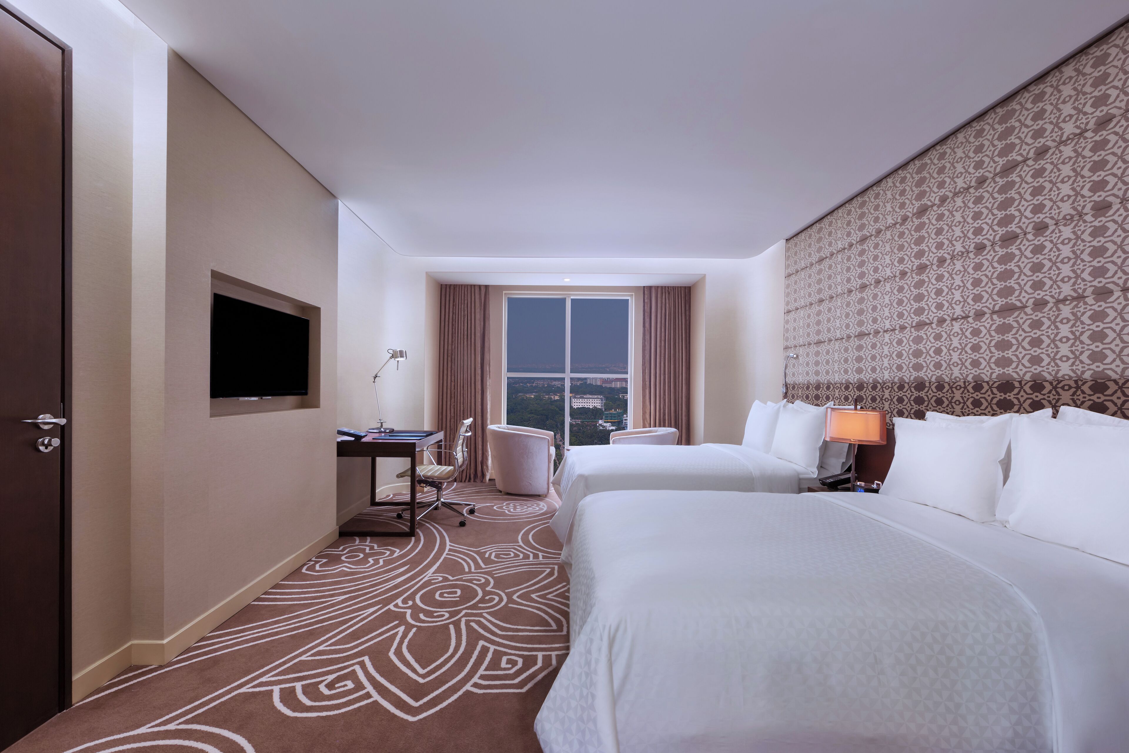 A hotel room with two beds, a desk, a chair, a TV, and a large window with a city view.