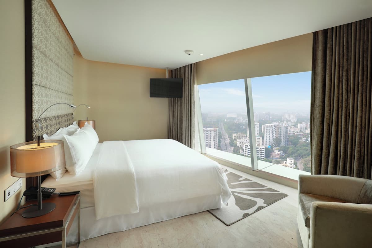Een hotelkamer met een groot bed, een bureau met een lamp, een aan de muur bevestigde televisie en uitzicht op de stad door grote ramen.