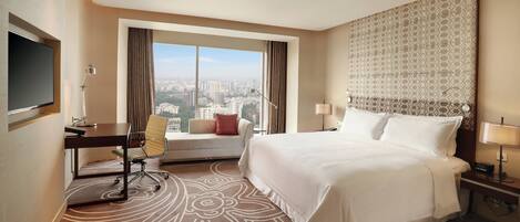 A hotel room with a large bed, a desk with a chair, a television, and a view of the city.