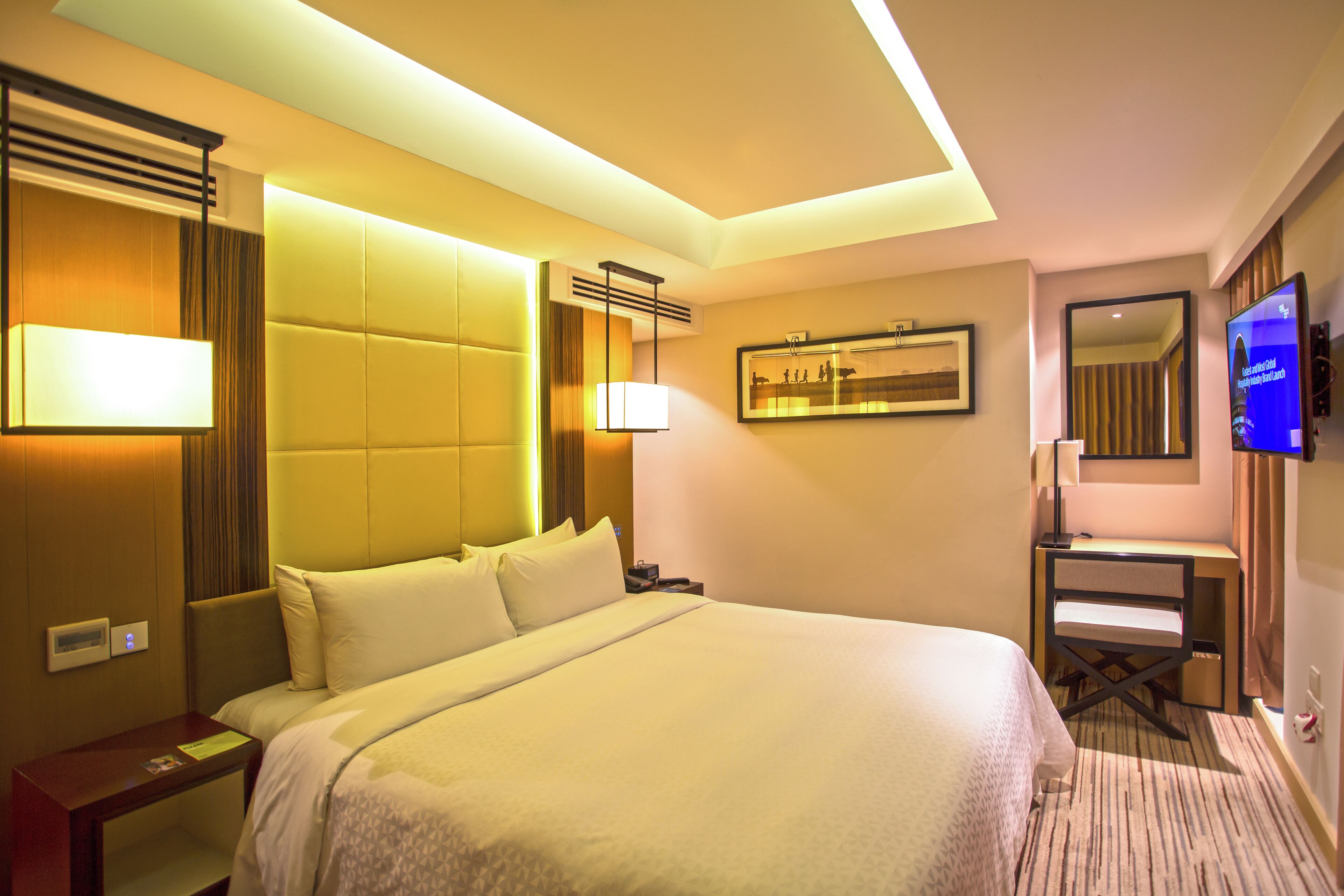 A hotel room with a large bed, bedside table, desk, chair, TV, and a framed picture on the wall.