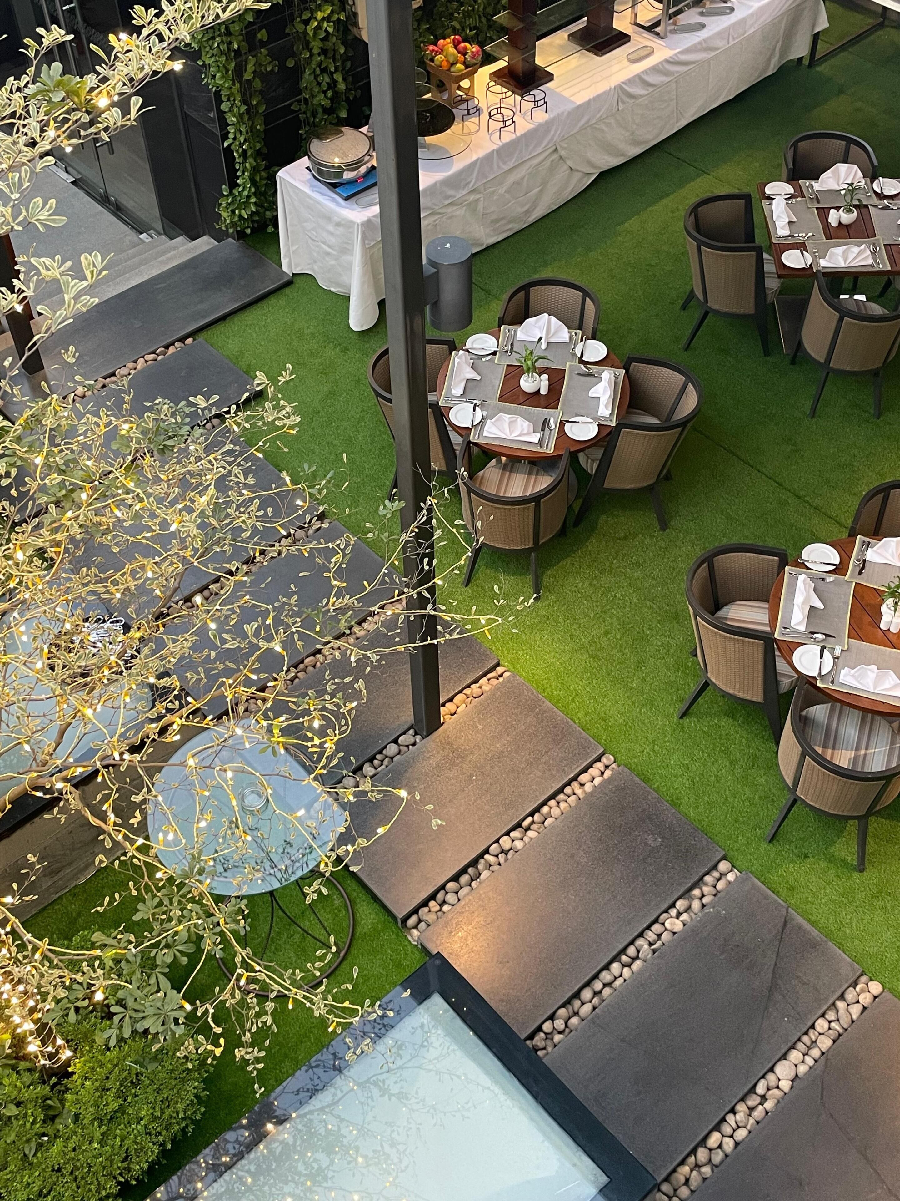 Un espacio de comedor al aire libre con varias mesas y sillas, rodeado de follaje e iluminación decorativa.