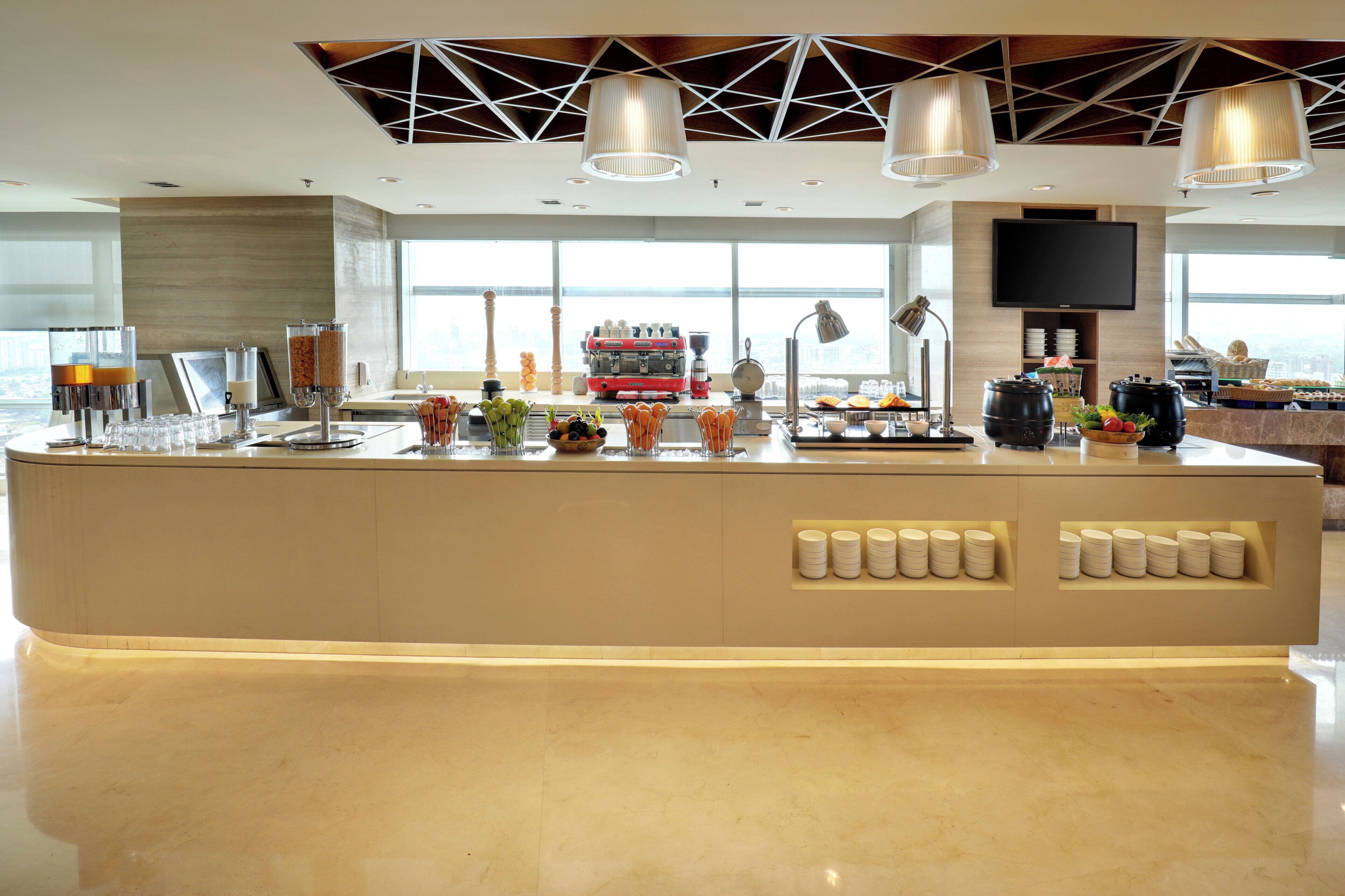 A modern kitchen area with a central island, equipped with various appliances and food items, and a large window providing a view of the cityscape.
