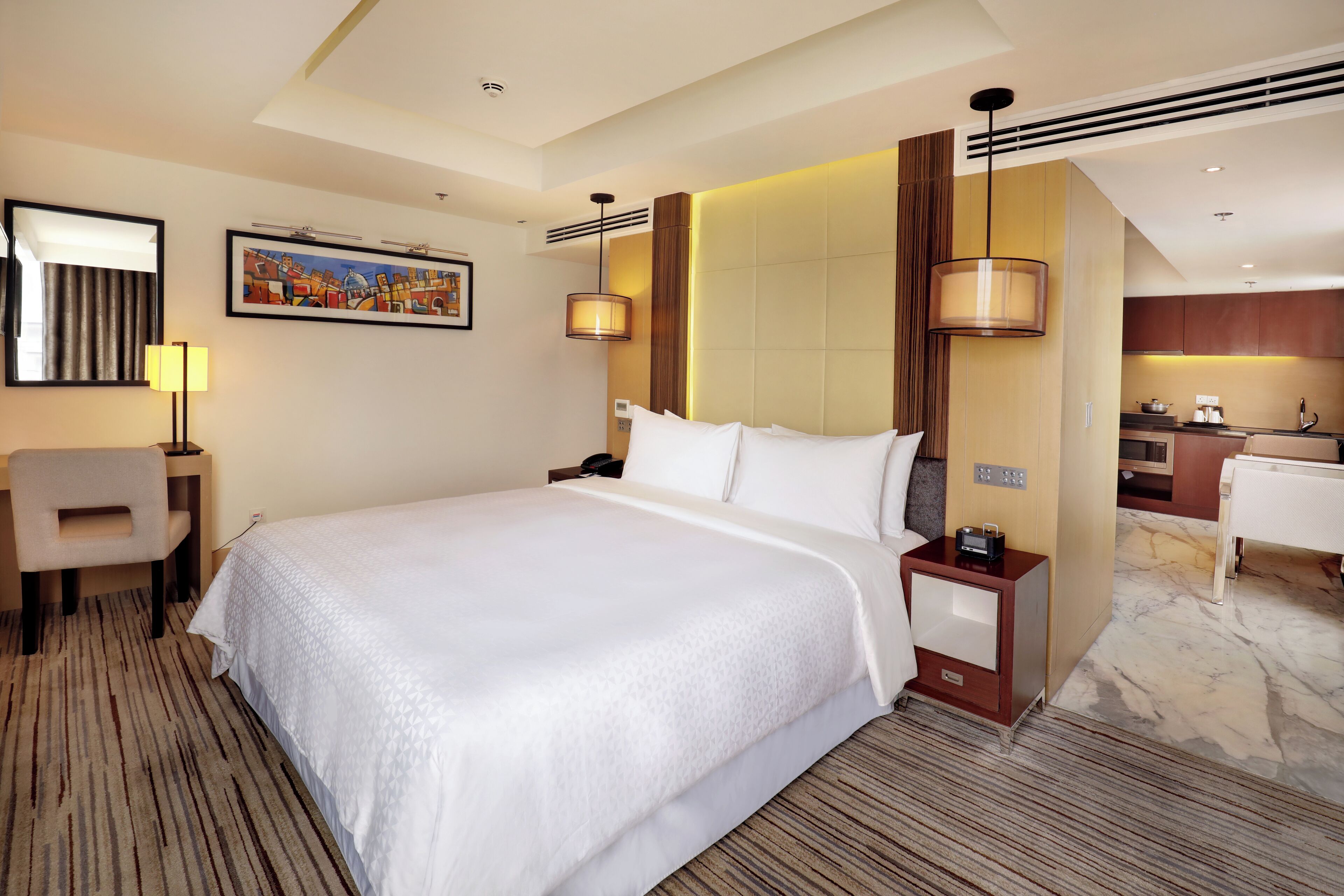 Quarto de hotel com uma cama grande, uma escrivaninha, uma cadeira, uma luminária de mesa, uma mesinha de cabeceira, um criado-mudo e uma televisão na parede, além de uma cozinha.