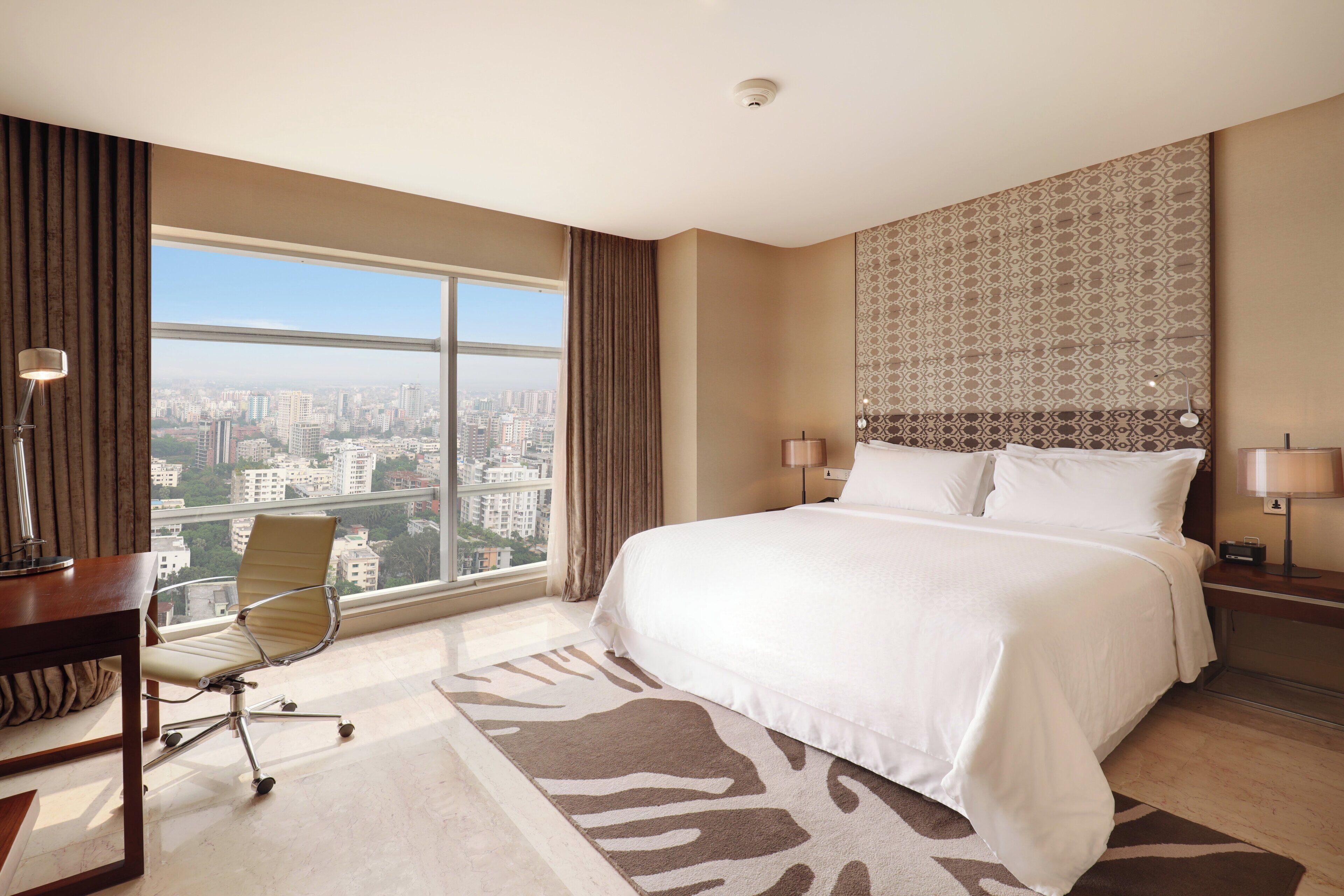 A hotel room with a large bed, a desk, a chair, and a view of the city.