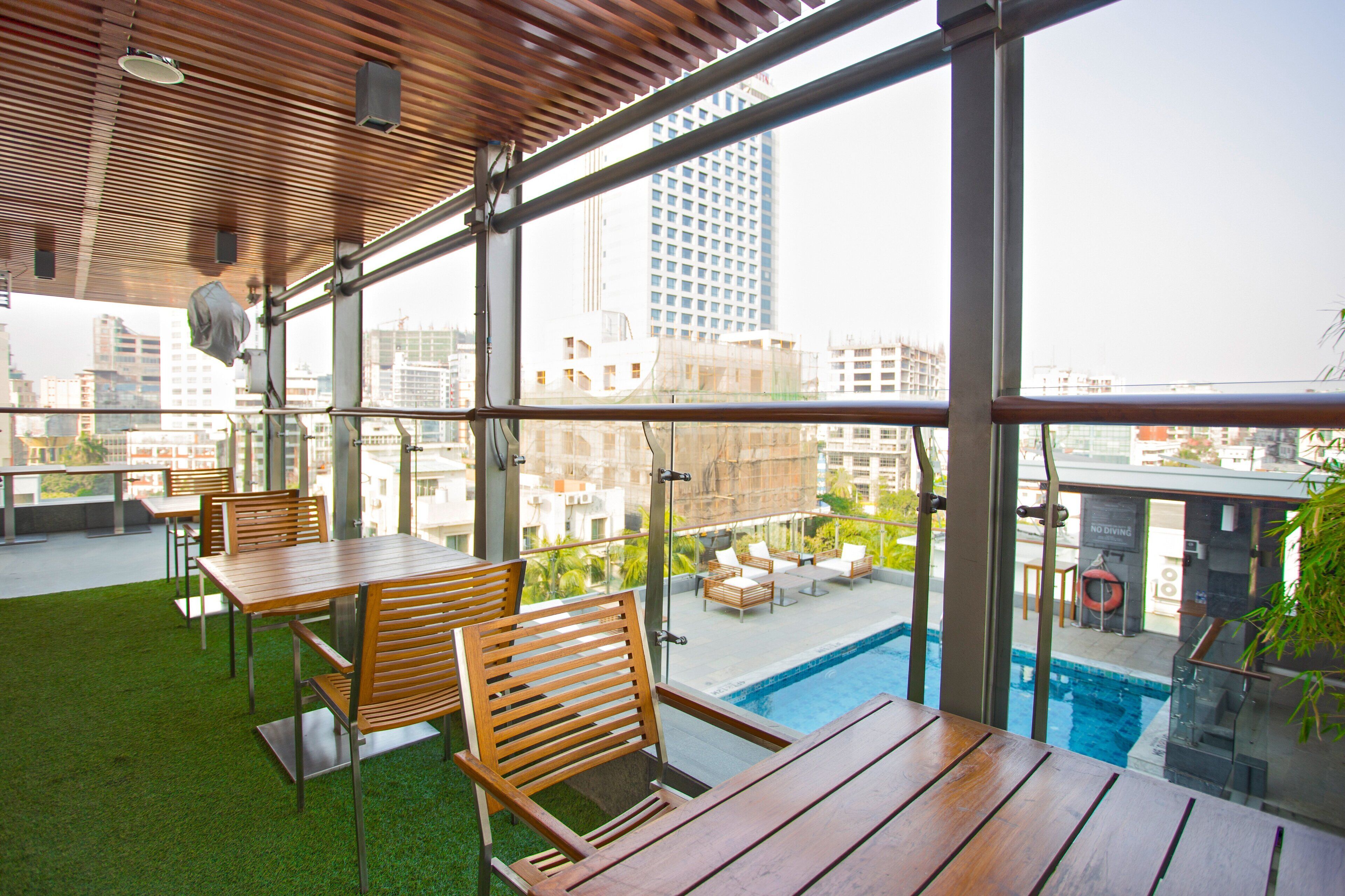 A rooftop terrace with wooden tables and chairs, a pool, and a view of the cityscape.