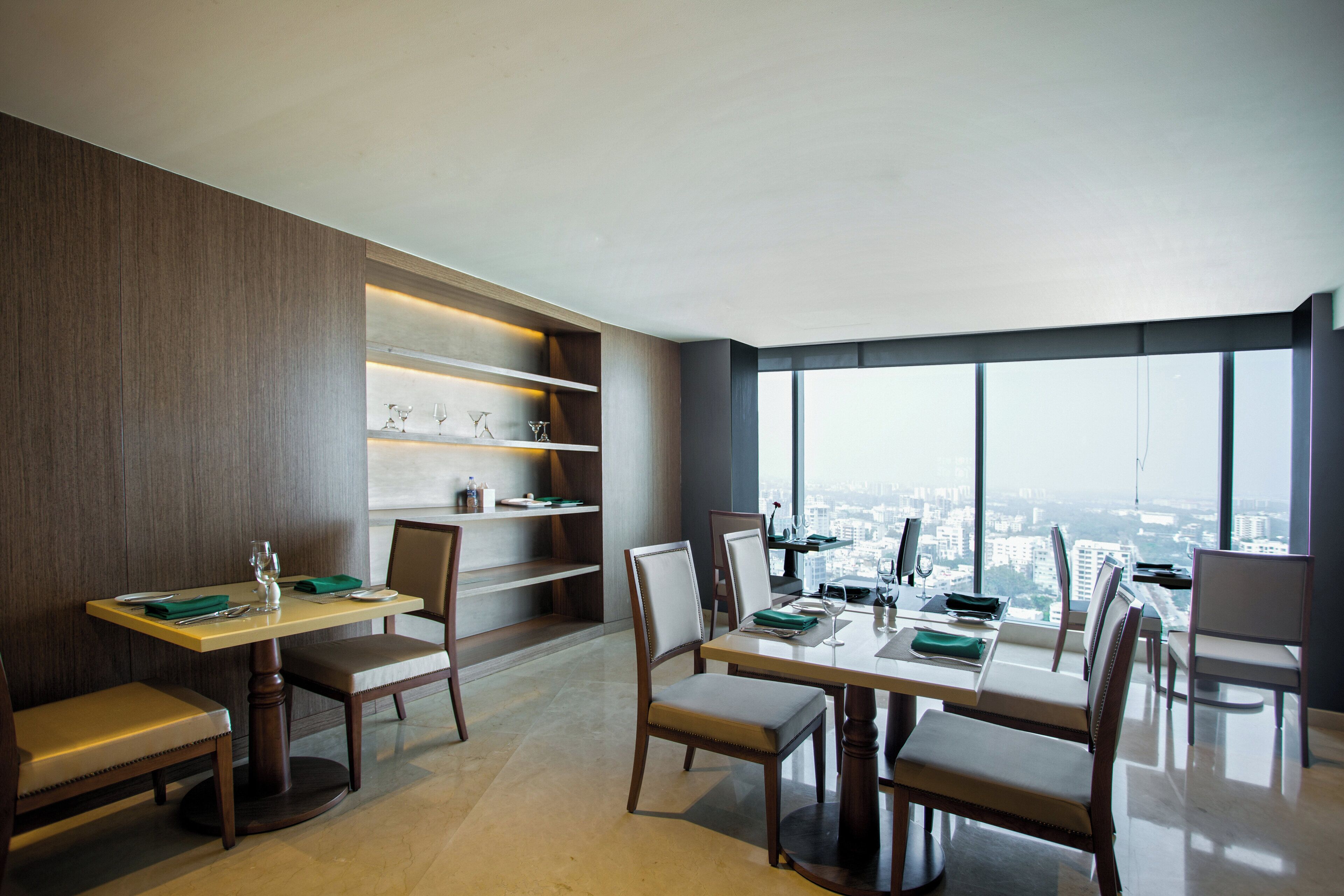 A modern dining area with a city view, featuring a table set for two, chairs, and a bookshelf.