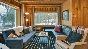 A cozy cabin interior with a wooden ceiling, a large window offering a view of trees, a blue sofa, a beige armchair, and a coffee table with a tray of pastries. - the Laurentides Private Log and Spa Escape (Rivière-Rouge)