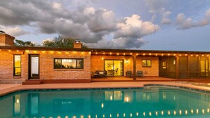 Una casa de ladrillo con piscina, mobiliario de exterior e iluminación con luces colgantes.