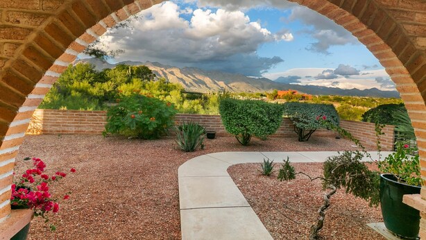 Un arco de ladrillo enmarca un jardín con un sendero de grava, diversas plantas y un paisaje montañoso al fondo.