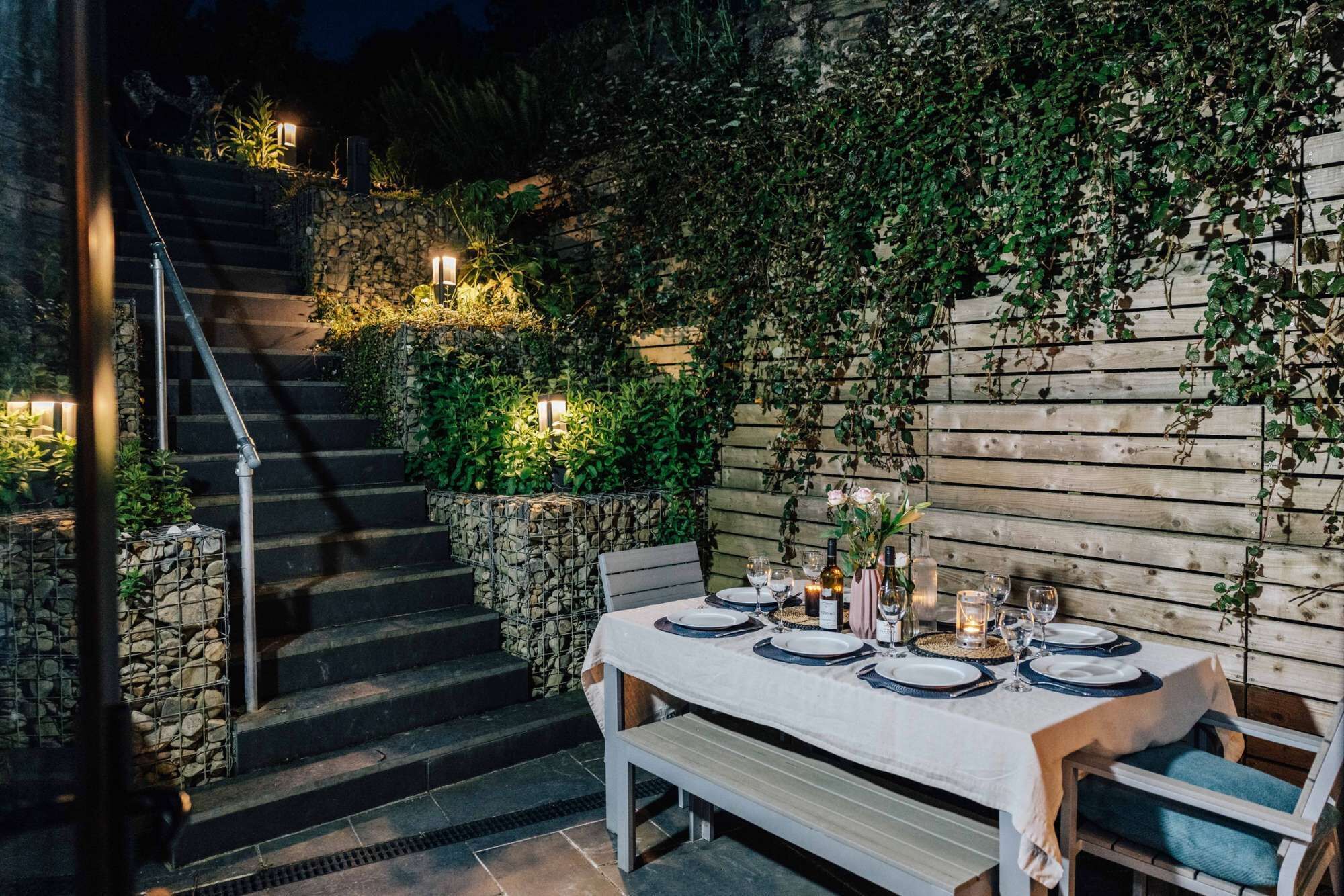 Un área de comedor al aire libre con una escalera de piedra, pared de madera y abundante vegetación.