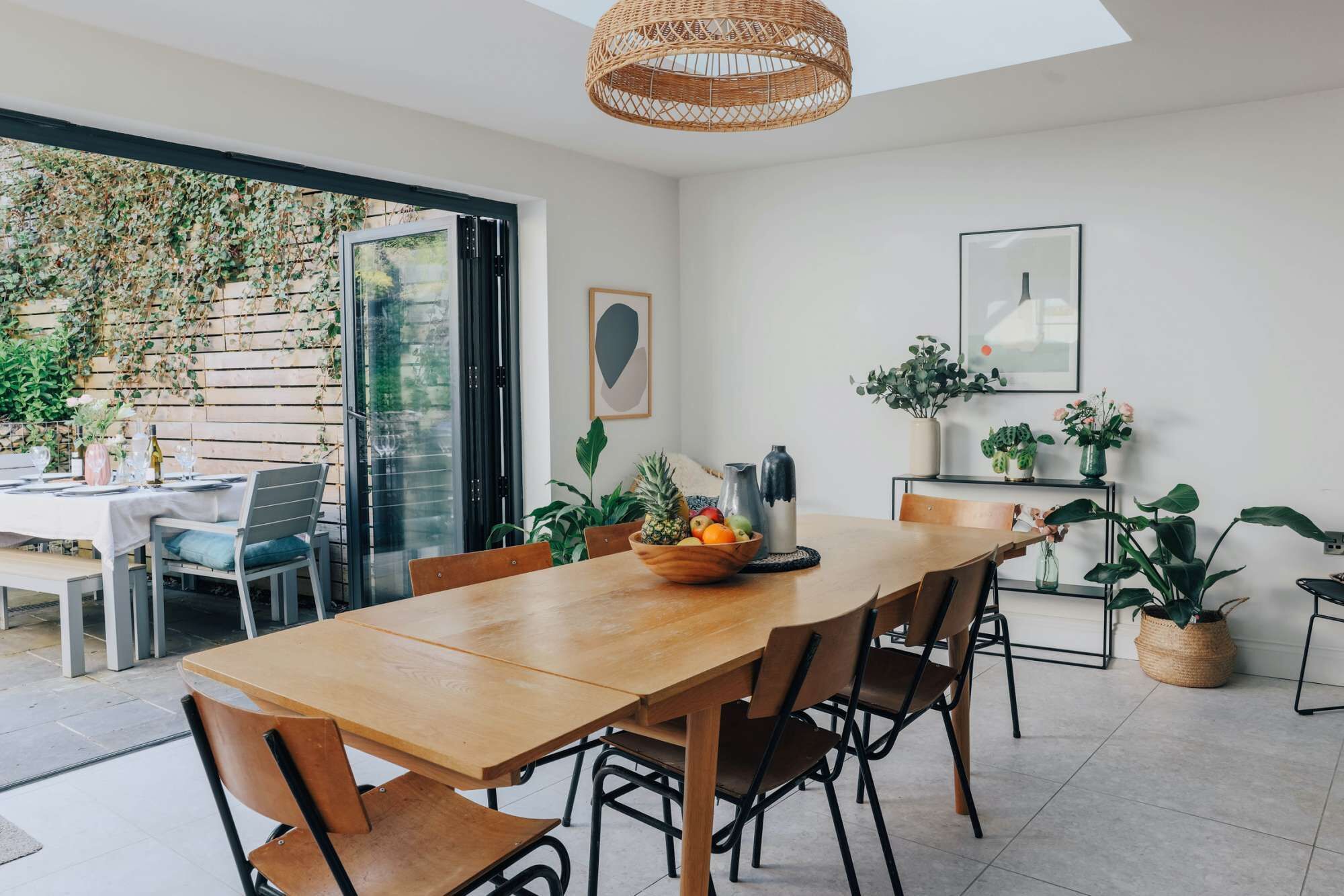 A dining area with a wooden table and chairs, a bowl of fruit, and potted plants.