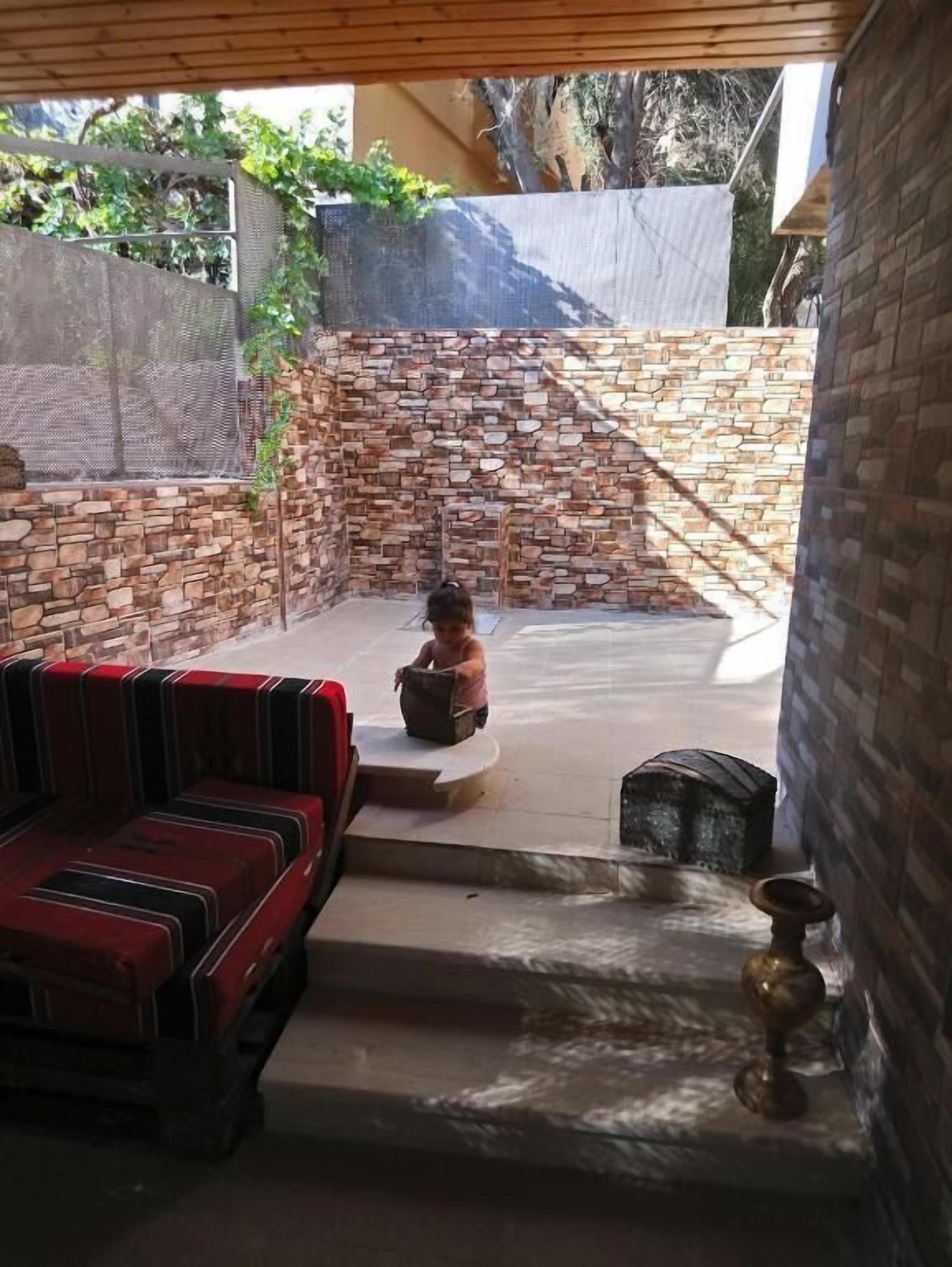 A covered outdoor area with a stone wall, a red and black striped sofa, and a child playing with a ball.