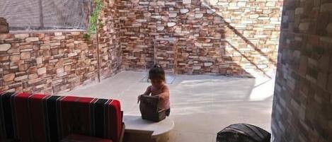 A covered outdoor area with a stone wall, a red and black striped sofa, and a child playing with a ball.