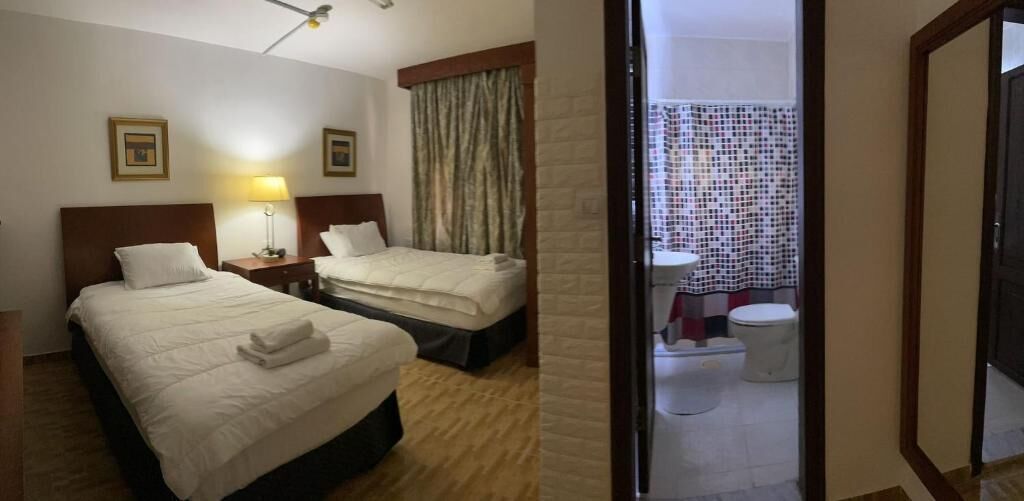Une chambre d’hôtel avec deux lits, une table de chevet avec une lampe, une fenêtre avec des rideaux et une salle de bain visible à travers une porte ouverte.